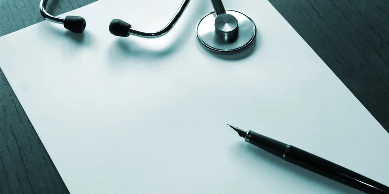 An overhead shot of a table with a stethoscope resting on a piece of paper beside a pen